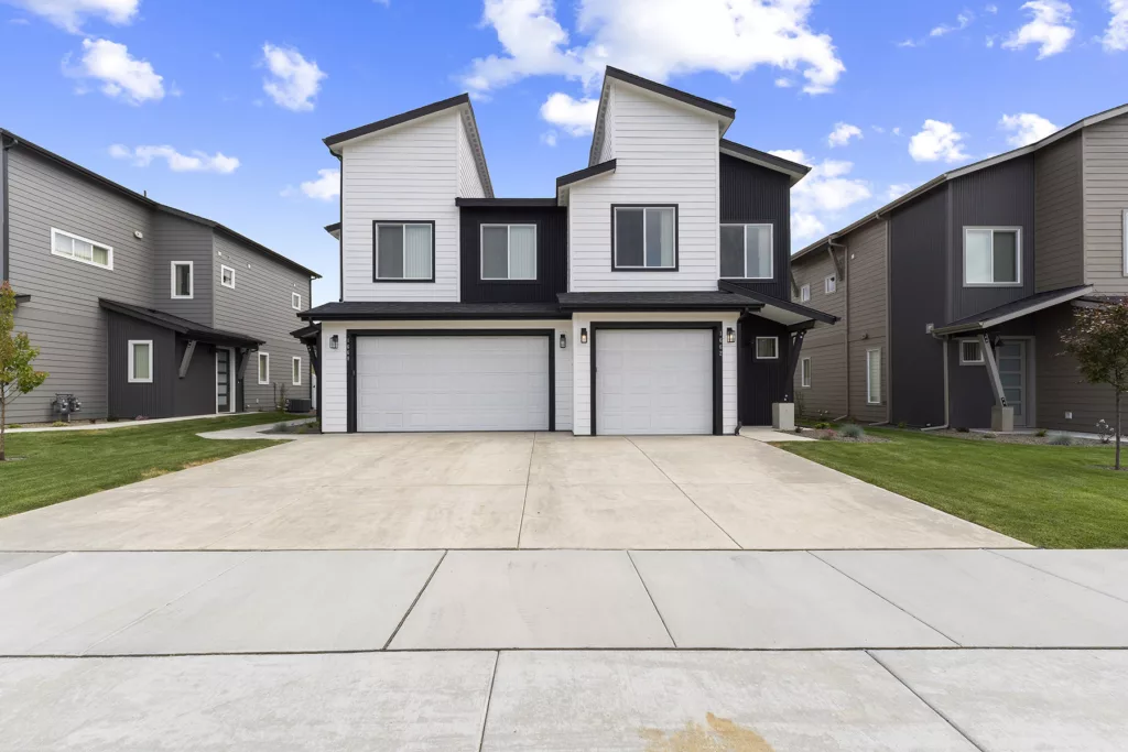 Modern duplex with a symmetrical facade, featuring white and dark siding, a shared double garage, and a neatly manicured lawn on a clear day.