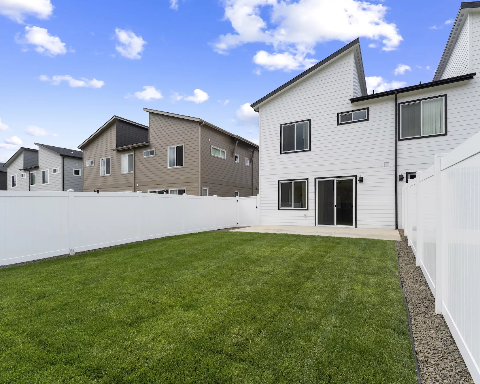Backyard of a modern two-story white house with a patio, green lawn, and white fence under a blue sky with clouds.