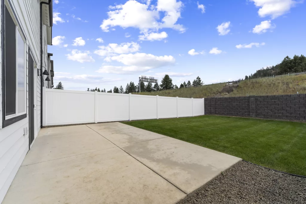 A backyard with a concrete patio, white vinyl fencing, green lawn, and a view of a hillside and trees.