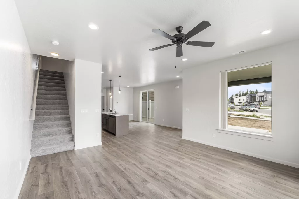 A spacious, empty room with wooden flooring, a ceiling fan, large windows, and a staircase leading to the upper floor.