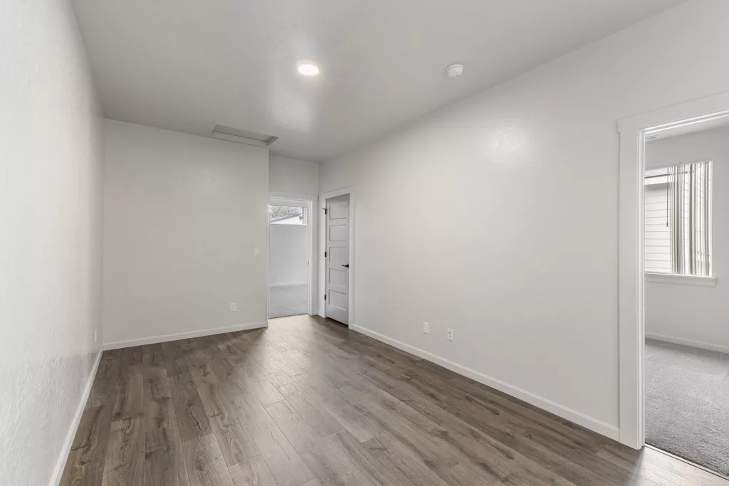 Empty room with white walls, hardwood floor, and a door leading to another room.