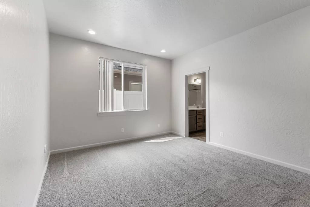 Empty room with light gray carpet, white walls, a window with vertical blinds, and an open doorway leading to another room.