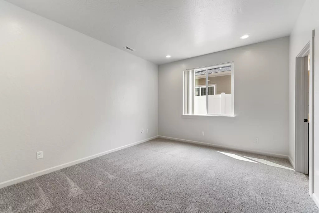 An empty room with beige carpet, white walls, a window with vertical blinds, and recessed ceiling lights.