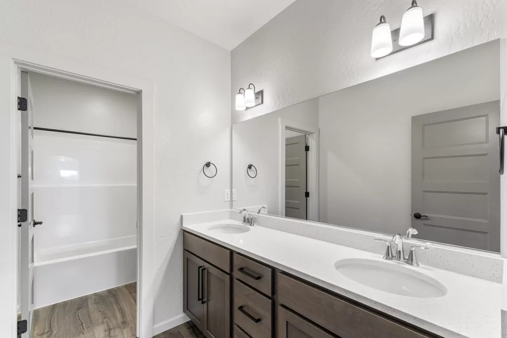 Modern bathroom with double sinks, dark wood cabinets, large mirror, and adjacent bathtub with white walls and wood flooring.