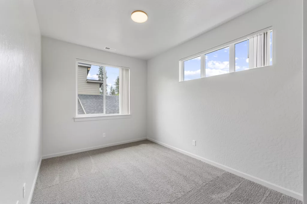 Empty room with white walls, gray carpet, and two windows allowing natural light in.