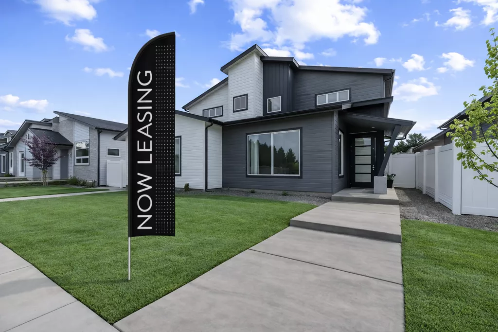 Modern gray and white house with a "Now Leasing" flag on a manicured lawn under a partly cloudy sky.